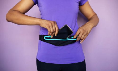 Mid section shot of a fitness woman in sports wear putting her smart phone into waist bag on purple background.